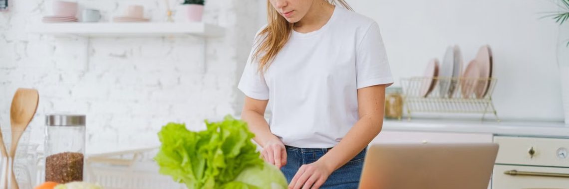 Woman in white t-shirt preparing fresh vegetables in a bright kitchen with a laptop.
